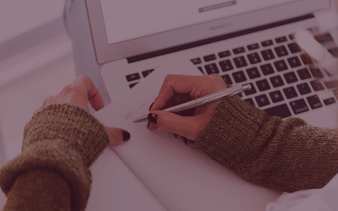 Aufgeklappter Laptop und Hände, die eine Hand hält einen Stift und schreibt in ein Notizheft